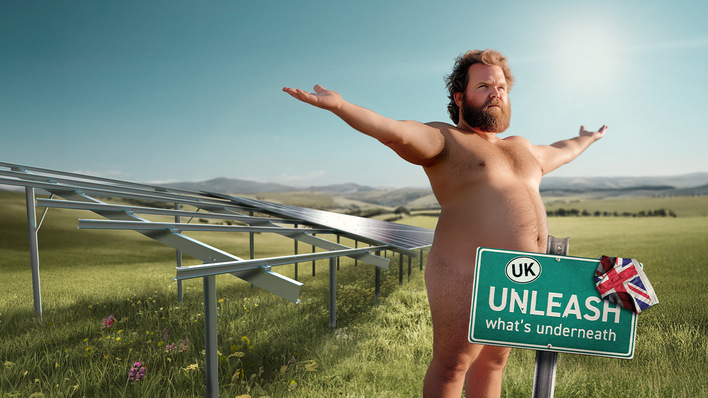 Ein nackter Mann steht mit ausgestreckten Armen neben Solarmodulen auf einem Feld, neben einem Schild mit der Aufschrift „UK UNLEASH“. - © PMT 2025
