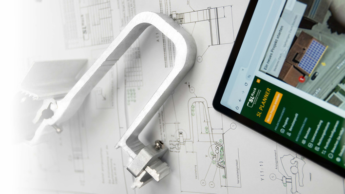 A metal clamp, technical drawings, and a tablet displaying a planning website are on a white surface. - © SL Rack SL Rack offer digital tools for real-life applications.